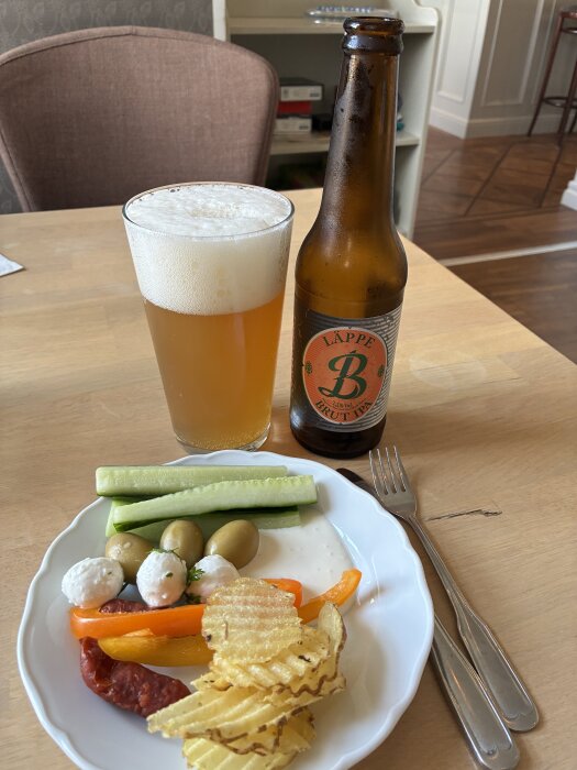 Ett glas öl med mycket skum, en flaska öl märkt "Läppe Brut IPA" och en tallrik med snacks inklusive chips, grönsaker och oliver.
