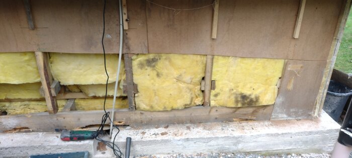 En vägg med gult isoleringsmaterial, delvis borttagen panel och ruttnande träsyll, redo för utbyte vid ett renoveringsprojekt.