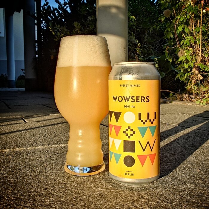 Ett glas öl bredvid en burk med etiketten "Wowsers DDH IPA" från Berlin, stående utomhus i solnedgången.