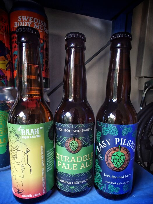 Tre ölflaskor på hylla, etiketter med “BAAH”, “Citradelic Pale Ale” och “Easy Pilsner”, samt burk med texten “Swedish Body Music” i bakgrunden.