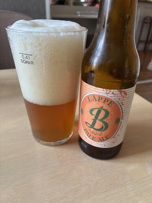 Ett glas med Pale Ale och mycket skum bredvid en flaska med etiketten "Läppe B Pale Ale", placerat på ett träbord.