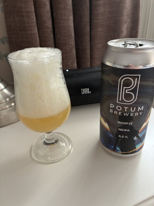 Ett glas öl med mycket skum bredvid en burk Potum Brewery Whip It NEIPA och en JBL-högtalare i bakgrunden.