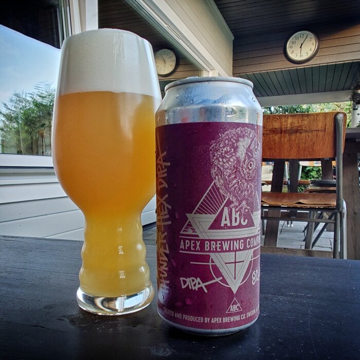 Ett glas med skummande öl bredvid en burk Apex Brewing Company öl på ett bord, inomhus med klockor på väggen i bakgrunden.
