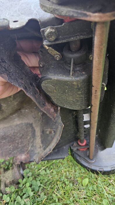 Gräsklipparens koppling snurrar fritt kring axeln, med remmen synligt lös och potentiellt saknade skruvar. En hand håller i komponenten.