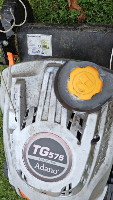 Gräsklippare TG575 med orange oljelock och märkning som visar 96 dB ljudnivå. Användaren har problem med kopplingen och lös rem.