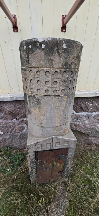 Stenstruktur cirka 1,5m hög med runda former och väderpåverkad yta, stående vid en mur med dörr och växtlighet runtom.