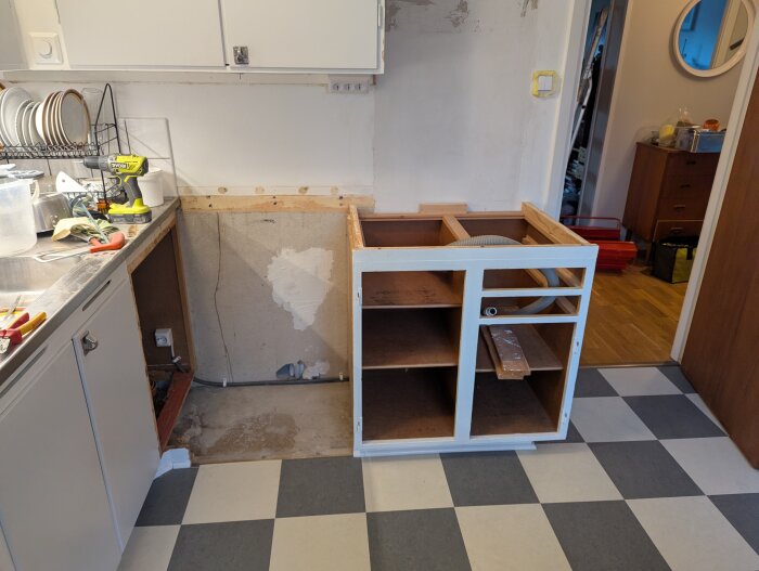 Under konstruktion av kök, exponerat skåp mot rutigt golv, synlig köksbänk och verktyg, vägg och betonggolv i renoveringsprocess.