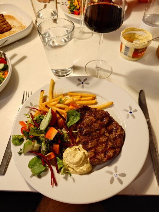 Grillad entrecote med pommes frites, bearnaisesås och sallad på tallrik, serverad med rödvin och vatten på ett matbord.