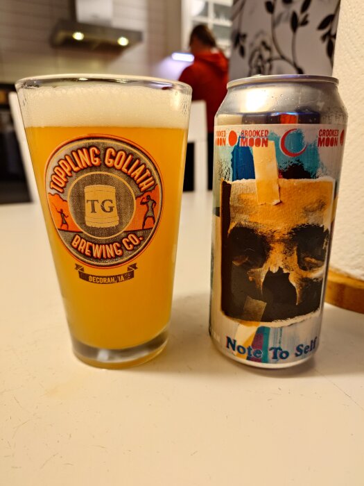 Glas med öl från Toppling Goliath och en burk Crooked Moon öl "Note To Self" med dödskallemotiv på ett bord.