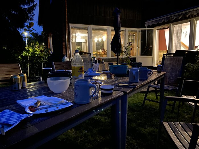 Kvällsmiljö med uteservering. Ett bord med blandade drycker och matrester, omgiven av trädgård och hus med tända fönsterlampor.