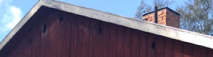 Fasaden på ett hus med flera stora hål under vindskivan, synlig plåttak och skorsten i bakgrunden.