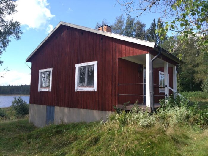 Rött hus med vita detaljer, beläget på landet vid en sjö. Stora hål syns i fasaden nedanför takskägget.