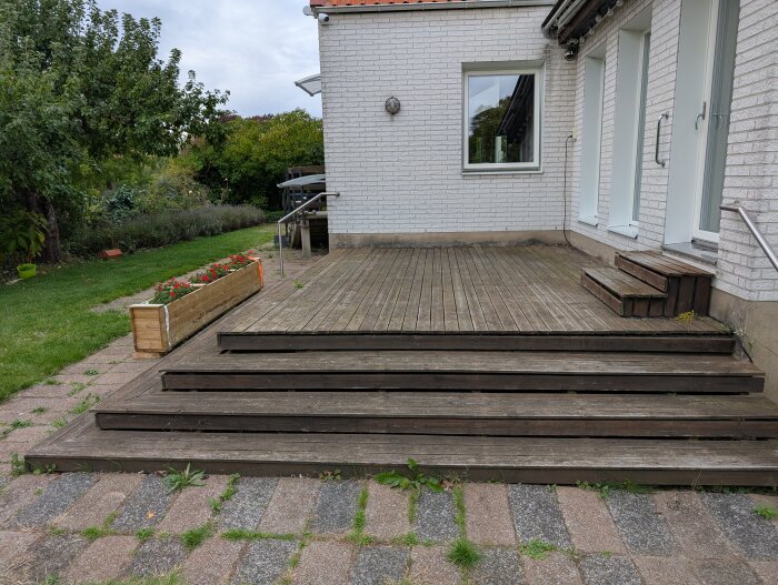 Altan med trappsteg i trä intill vit tegelvägg, gräsmatta och blomlåda fylld med röda blommor, fotograferad vid ett hus.