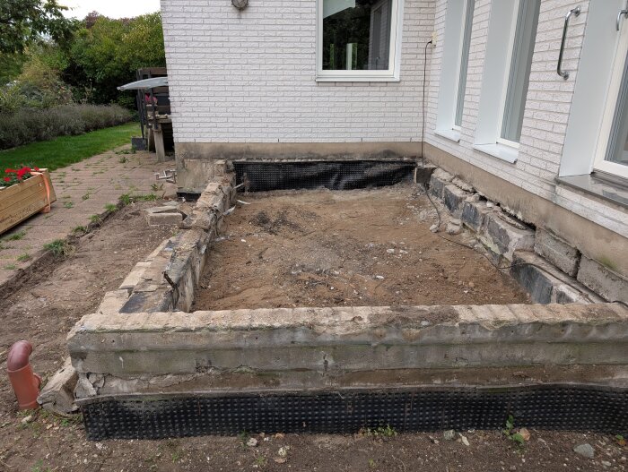 Borttagen altan vid vit putsfasad med synliga grundmurar och jord. Trädgård med gångväg och blomlåda i bakgrunden.