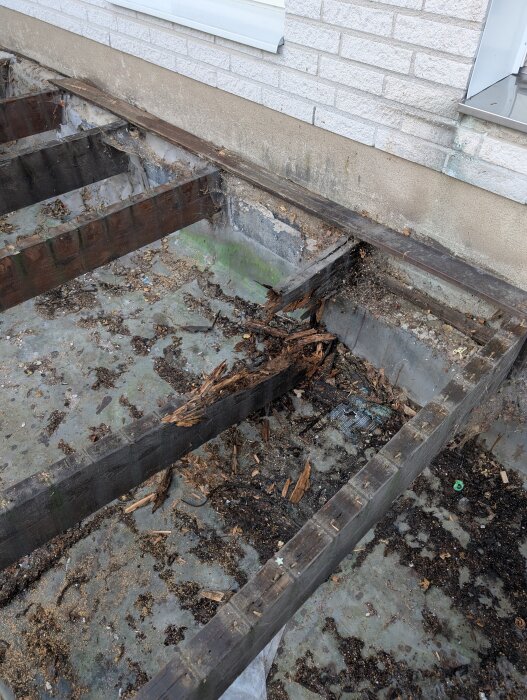 Riven altan med synliga bjälklag, där en balk har ruttnat av helt. Rester av trä och skräp ligger på marken intill husfasaden.