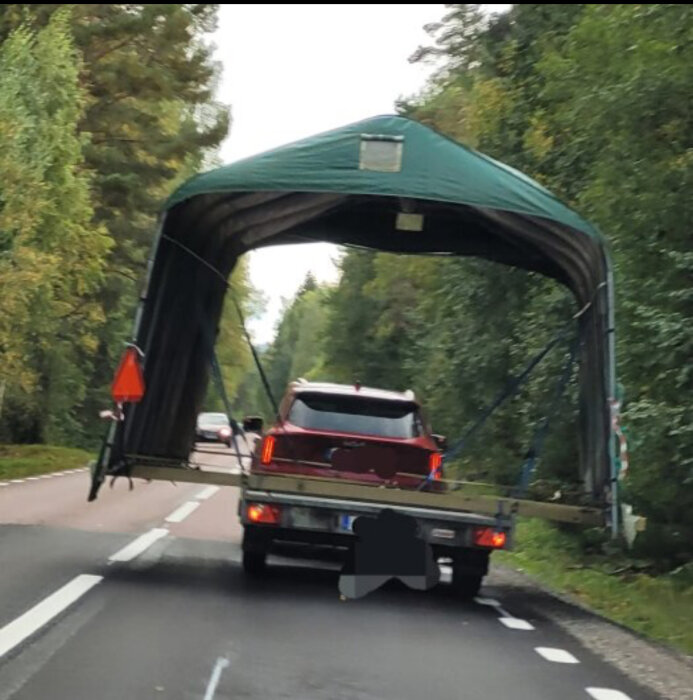 En röd bil som kör på en landsväg, lastar en stor, grön tältliknande struktur ovanpå en släpvagn, sedd bakifrån.