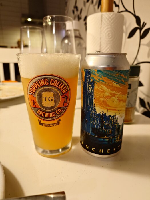 Öl från Northern Monk med burkdesign av stadssilhuett, serverad i ett glas från Toppling Goliath Brewing Co., på ett köksbord.