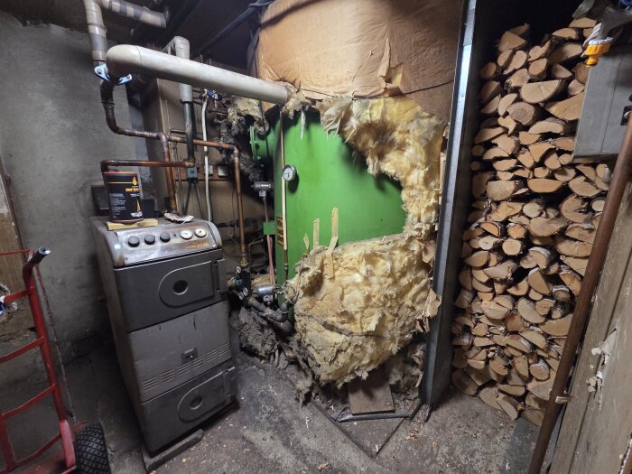 Pannrum med vedpanna och rörelement, isolering skadad, vedstapel till höger.