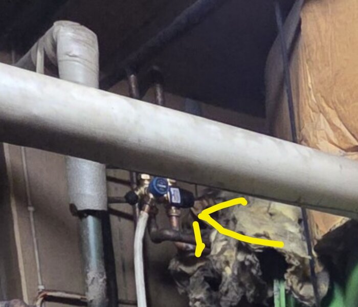 Gul pil pekar på termostat/blandningsventil för tappvarmvatten i ett rörsystem, omgiven av rör och isolering.