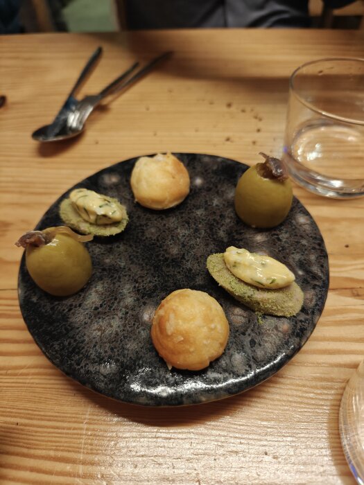 Trevlig avsmakning med små rätter på en svart tallrik, troligen tapas, bredvid ett glas vatten och bestick.