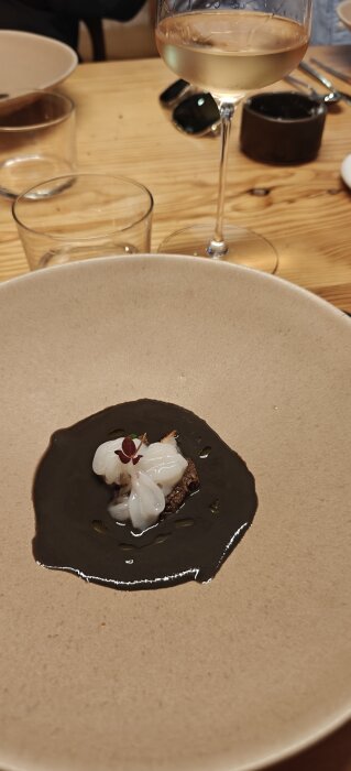 En elegant rätt på en tallrik med svart sås och vit garnering, bredvid ett glas vitt vin på ett träbord.