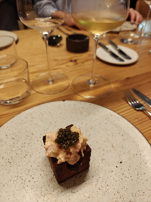 Bröd med laxkräm och caviar på tallrik, omgiven av vinglas på ett träbord.