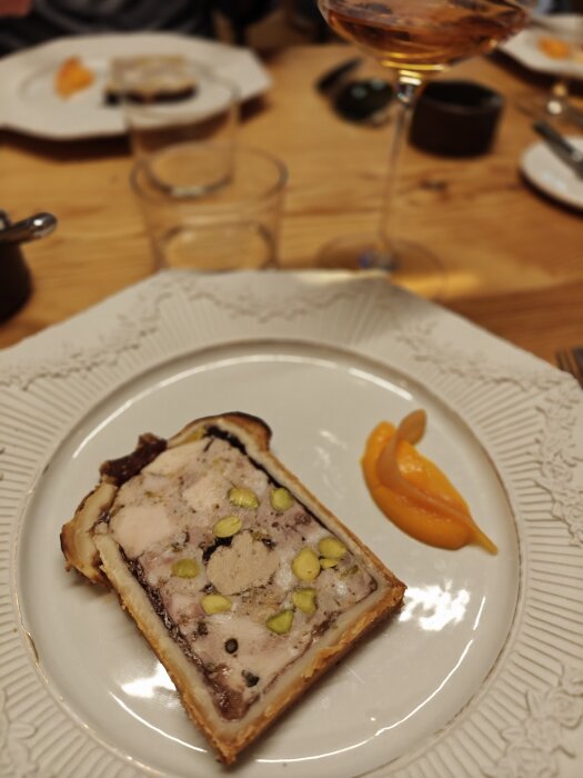 En elegant serverad pâté med pistagenötter på en tallrik, garnerad med en klick orange puré och ett glas vin i bakgrunden.