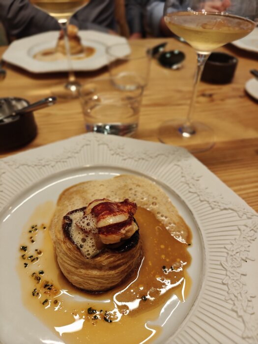 Plate with pastry topped with seafood and sauce, wine glasses and table setting in background.