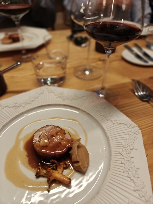 Tallrik med köttbit, sås, kantareller och puré, omgiven av vinglas och bestick på ett dukat bord.