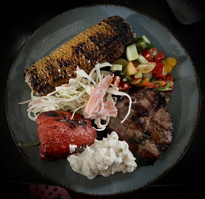 Grillad fläskkarré med grillad majs, ostfylld paprika, kålsallad, potatissallad och sommarsallad på tallrik.
