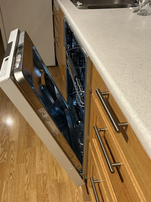 Öppen diskmaskin i kök med träskåp, där luckan syns dra ut från maskinen men inte i nivå med bänkskivan.