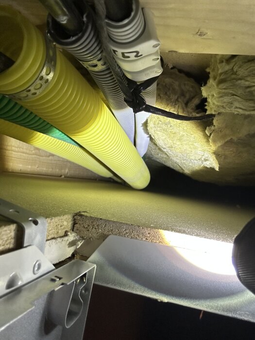 Elrör och isolering syns under entrén, med gula och gröna rör samt mineralull. Inga synliga tecken på mögel, men det finns en dålig lukt.