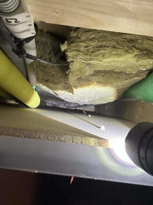 Rör och isolering i ett hus, sett från insidan. Elrör i olika färger och en inspektionslucka. Ficklampa lyser upp området.