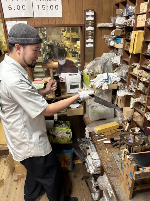 En man arbetar i en verkstad, håller en kniv och hammare, omgiven av verktyg och material.