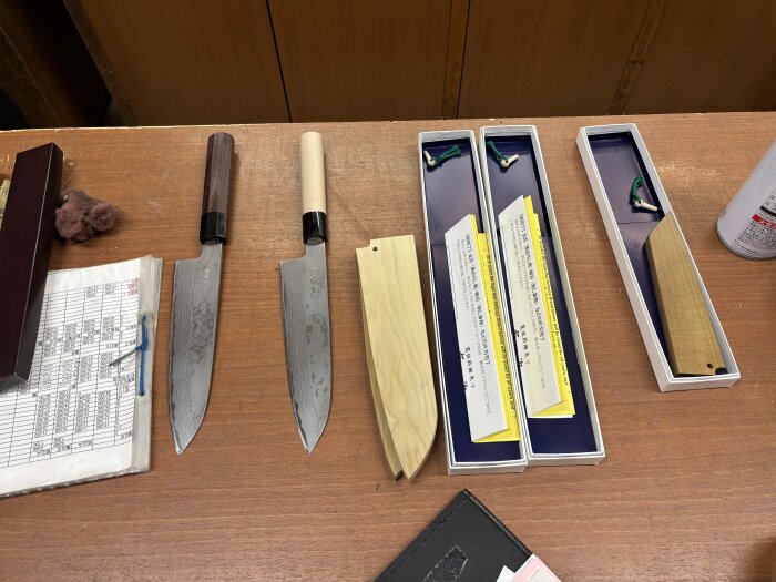 Två japanska knivar med damaskusmönstrade blad och trähandtag ligger bredvid sina förpackningar, dokument och en knivslida på ett bord.