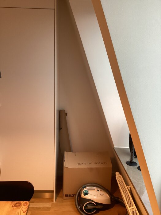 Snedställt trekantigt utrymme i kök med kartong och dammsugare på golvet, mot en sned yttervägg.