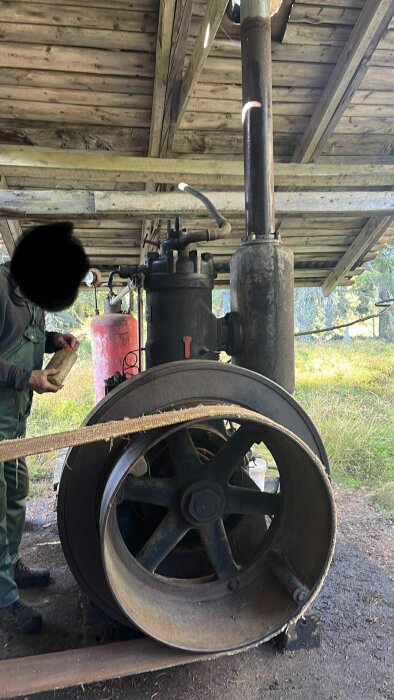 Tändkulemotor under tak, stora hjul, en person håller något i handen, grönt gräs och träd i bakgrunden.
