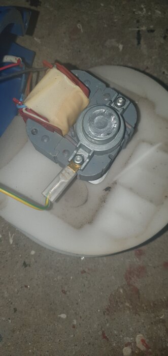 Defekt fläktmotor från en Separett Villa 9000 med synliga ledningar och monteringsdetaljer, placerad på ett smutsigt underlag.