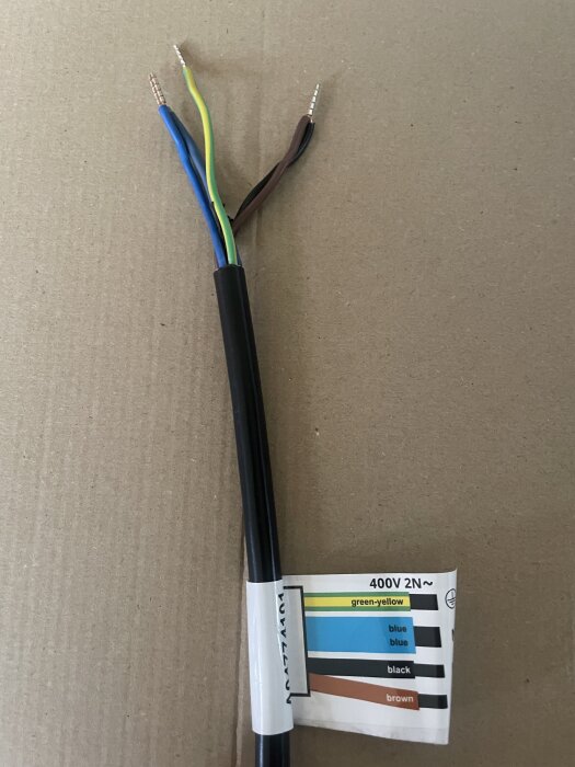 Kabel med fyra ledare i olika färger; brun, svart, blå och grön-gul, visas mot en kartongbakgrund med märkningsetikett för installation.