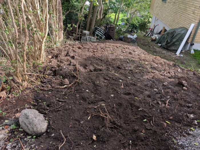 Upphöjd markyta med jord och fyllnadsmassor omgiven av buskar och träd, med en husvägg och trädgårdsföremål i bakgrunden.