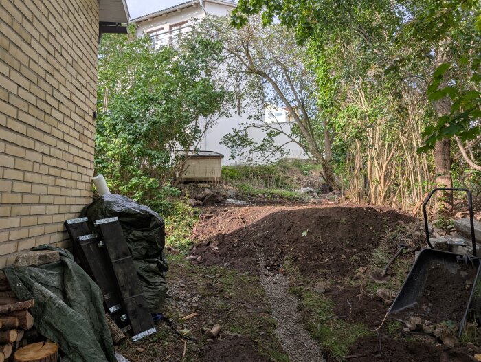 Upphöjd jordig yta med träd i bakgrunden och byggmaterial på marken, bredvid ett gult tegelhus. En skottkärra syns till höger.