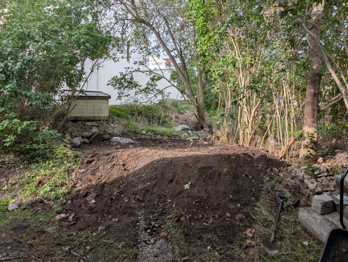 Upphöjd jordyta i en trädgård med buskar och träd i bakgrunden, en spade och stenar syns i förgrunden.