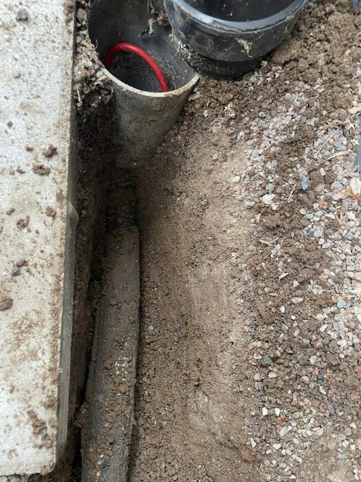 En grävd markyta med synlig svart slang och en röd mindre slang i ett avloppsrör, nära husfasad.
