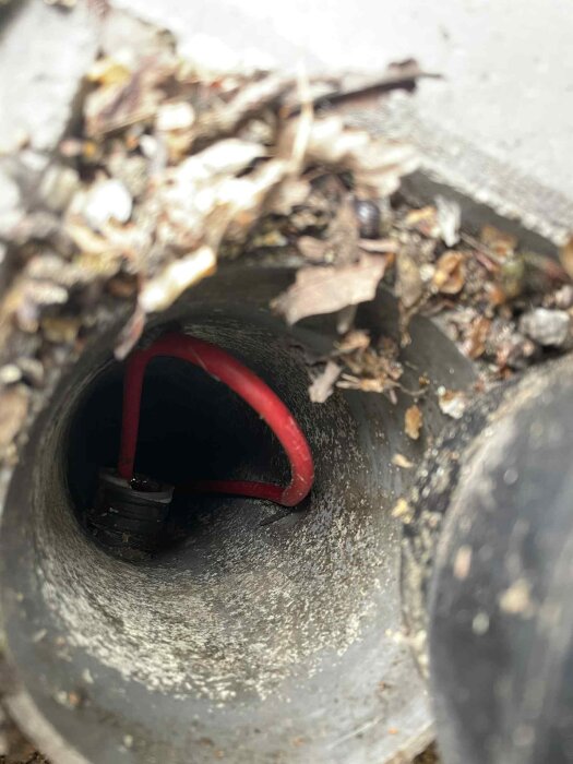 En bild av ett öppet rör nära en husfasad med en röd slang inuti, omgiven av jord och löv. Det ser ut som slangen kan ha blivit lagad.