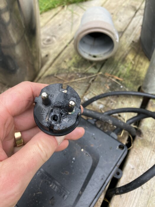 Bränd och delad elkontakt från en pump, hålls i handen utomhus på en träyta, med kablar och rör i bakgrunden.
