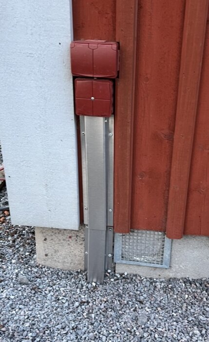 Rött eluttag på vägg med rostfritt kabelskydd som går ner till grus. Vägg i rött trä och vit puts.