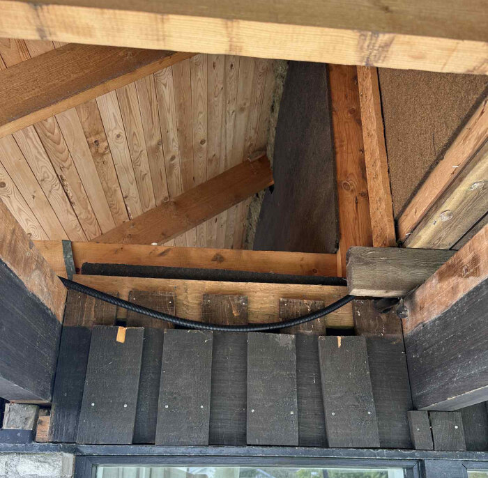 Trävägg med exponerat bjälklag och takstolar, svart kabel och metallremsa ovanför en glasdörr i del av kattvind i 1,5-planshusets övervåning.