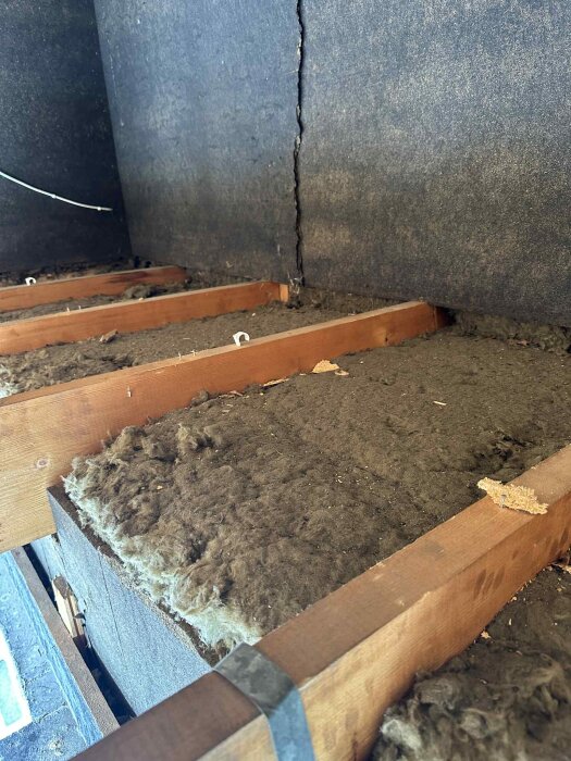 Isolering mellan bjälkar på en ovanvåning, med en metallremsa ovanpå. Vägg med skarv i bakgrunden.