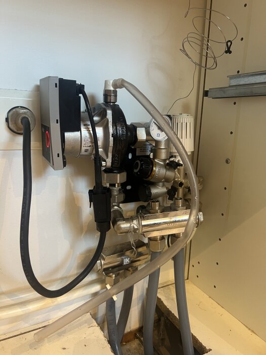 Golvvärmeshunt med anslutna rör och elektroniska komponenter installerad i ett skåp, synlig märkning "LK" och kabeldragning.