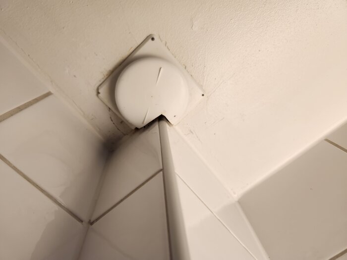 Vit ventilationsfläkt monterad i ett hörn av ett rum, omgiven av vita kakelplattor på väggen.